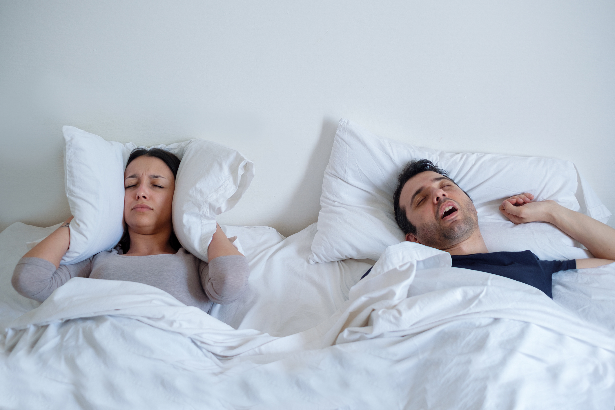 Couple in bed with the woman holding a pillow over her ears and a man snoring loudly.