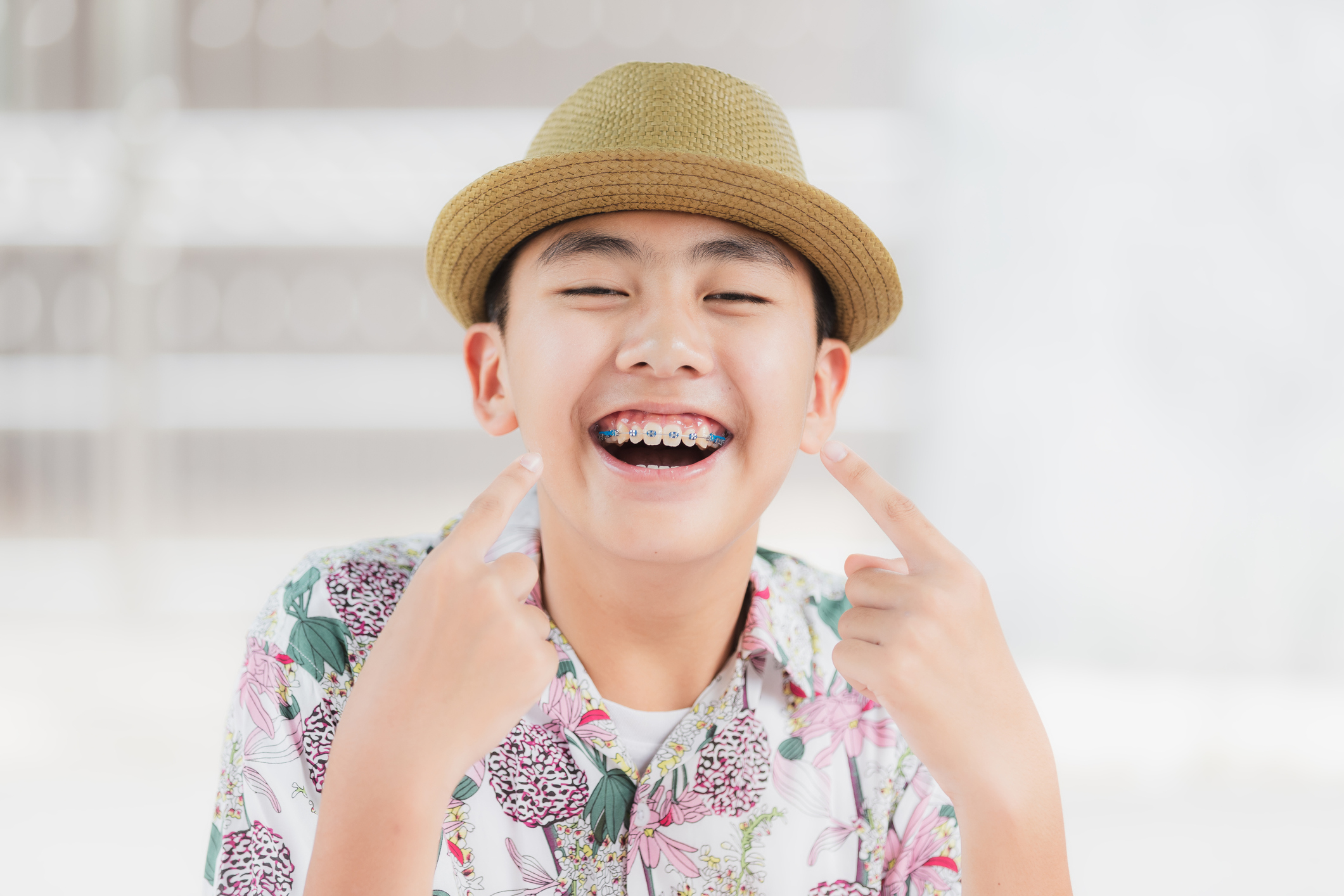 Young boy wearing a hat smiling pointing at his teeth which have braces.