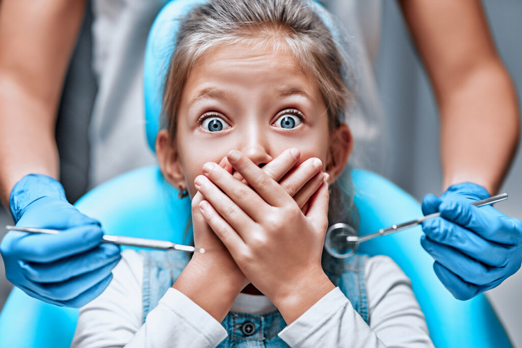 Close up view of a little girl looking scared and terrified screaming covering her mouth from the dentists with medical tools. 