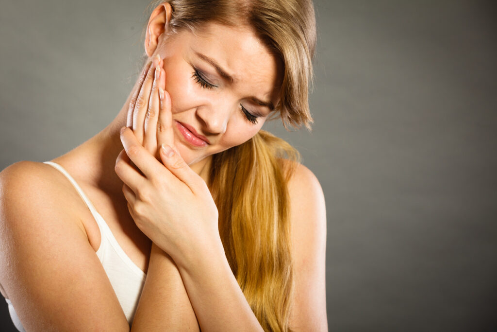 Young woman achy girl suffering from terrible tooth pain, touching pressing her cheek by hand palm.