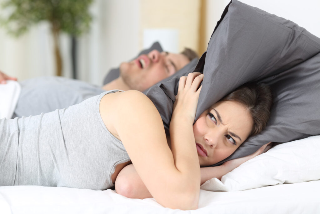 woman in bed holding pillow over her ears with husband snoring in background